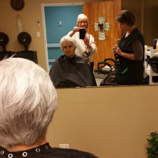 A senior woman receives a haircut at the salon, while stylists assist her in front of a mirror.
