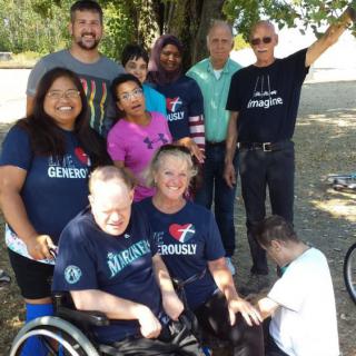 A diverse group of people gathered outdoors under a tree, with some wearing matching t-shirts and one person in a wheelchair.