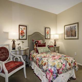 A cozy bedroom features a single bed with floral bedding, accompanied by nightstands and lamps on either side. A decorative chair and framed art complete the inviting ambiance.