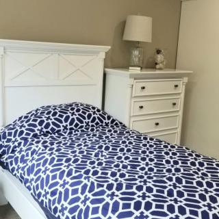 A neatly arranged bedroom featuring a white bed with patterned bedding and a matching dresser with a lamp.