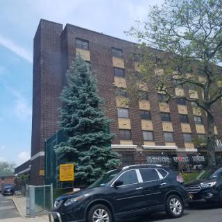 A brick building with boarded windows, accompanied by parked cars and a green tree in the foreground.