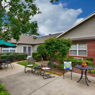 A cozy outdoor patio area with tables, chairs, and a sun umbrella, surrounded by greenery and adjacent to a building.