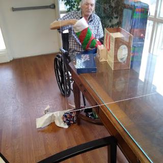 An elderly person in a wheelchair sits at a table surrounded by holiday gifts.