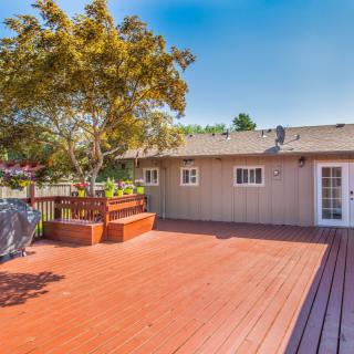 A spacious wooden deck connected to a house, featuring a tree and outdoor seating under a clear blue sky.