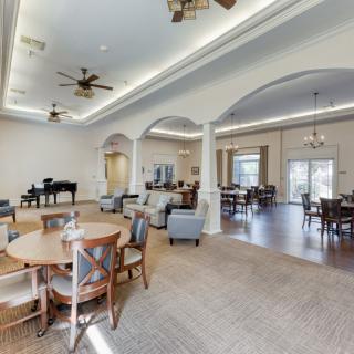 A spacious, elegantly designed dining area with round tables, chairs, a piano, and ceiling fans.