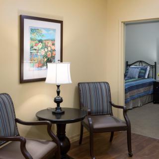 A cozy living area with two chairs, a round table, and a lamp, adjacent to a bedroom.