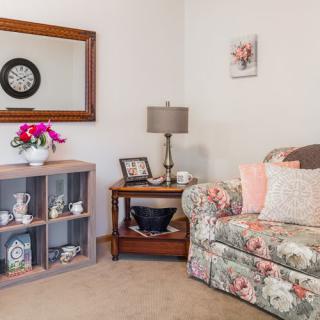 A cozy living room corner with a floral couch, decorative mirror, and shelving unit adorned with various decor items.