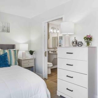 A cozy bedroom with a white dresser, bed, and nightstand, leading to an adjoining bathroom.