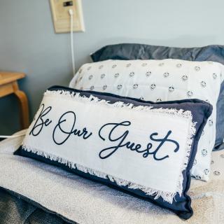 A cozy bed with a decorative pillow featuring the text 'Be Our Guest'.