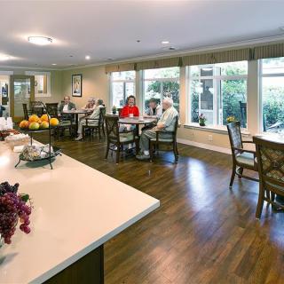 A communal dining space with tables and chairs, where several seniors are seated and enjoying a conversation.