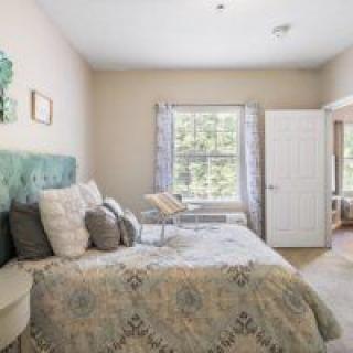 A cozy bedroom with a large bed covered in decorative pillows, a patterned blanket, and natural light streaming through the window.