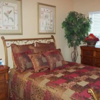 A cozy bedroom with a quilted bedspread and decorative pillows, featuring framed artwork and a potted plant.