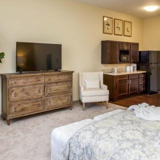A cozy bedroom with a television, dresser, and a small kitchenette.