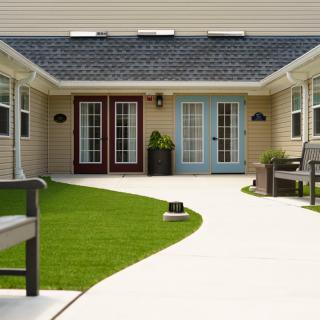 A neatly landscaped courtyard with benches, grass, and a smooth pathway leading to multiple doors of a building.