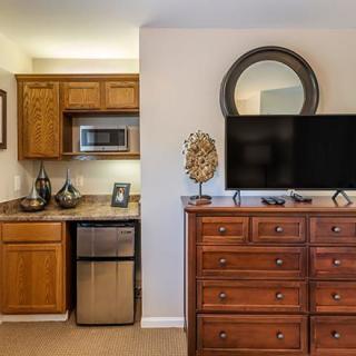 A cozy bedroom corner featuring a wooden dresser with a mounted TV and decorative mirror, alongside a kitchenette area with a microwave and mini fridge.
