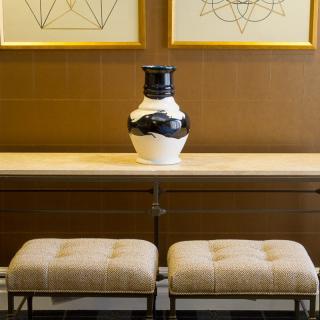 A modern interior featuring a console table with a black and white vase, flanked by two upholstered stools under framed geometric artwork.