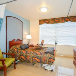 A well-lit hospital room with a single bed, chair, bedside lamp, and large window.