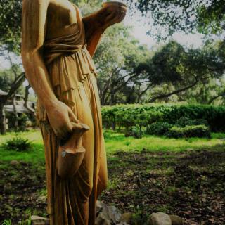 A classical statue in a serene garden setting surrounded by lush greenery and trees.