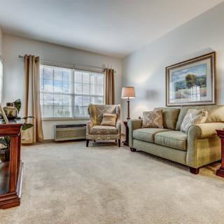 A cozy living room with a sofa, armchair, and soft carpet, complemented by a large window and decorative artwork.