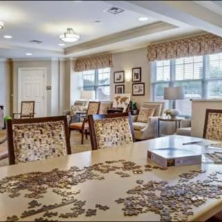 A cozy living room with a puzzle on the table, surrounded by comfortable seating and natural light from large windows.
