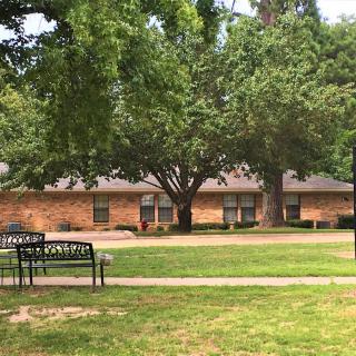 A brick building surrounded by lush green trees and a grassy lawn, with benches placed along the pathway.