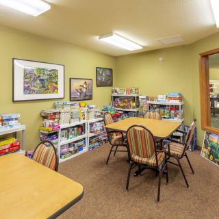 A cozy room with tables and chairs, surrounded by shelves filled with books and games, under bright lighting.