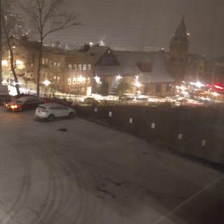 A serene winter night scene with snow blanketing the ground and lights illuminating the buildings.