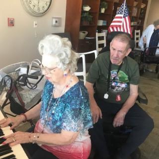 An elderly woman plays the piano while a man sits nearby, with a flag visible in the background.