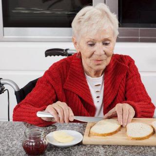 An elderly woman in a red sweater is preparing a meal, spreading butter on slices of bread at a kitchen counter.