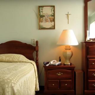 A cozy bedroom with a wooden bed, nightstand with a lamp, and a dresser with a mirror. A painting and cross adorn the wall.