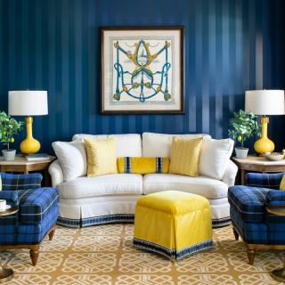 A stylish living room featuring a white sofa with yellow cushions, complemented by plaid blue chairs and a patterned rug. The space is highlighted by striped blue walls and bright yellow lamps.
