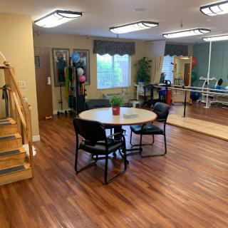 A bright rehabilitation space featuring a dining area with a table and chairs, exercise equipment, and wooden flooring.