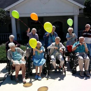 A joyful group of elderly individuals holding colorful balloons and sitting in wheelchairs outdoors.
