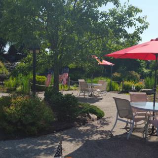 A sunny garden patio with chairs, tables, and red umbrellas surrounded by lush greenery.