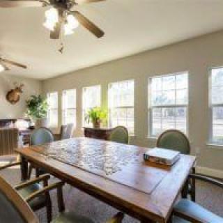 A well-lit dining room with a wooden table and chairs, featuring large windows and a ceiling fan.