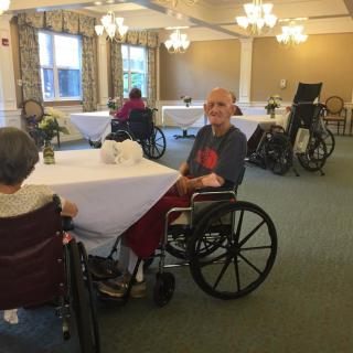 Elderly people in wheelchairs are sitting at tables in a nursing home dining area.