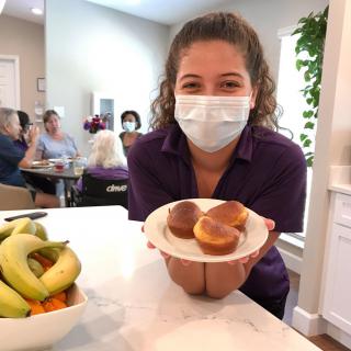 A woman wearing a mask is smiling and holding a plate of buns in a kitchen setting.