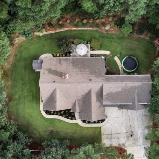 Aerial view of a house surrounded by trees, featuring a well-maintained lawn, a trampoline, a patio area, and a driveway.