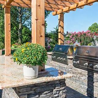 A beautifully designed outdoor kitchen featuring stone countertops and a pergola, complete with multiple grills and surrounded by lush greenery.