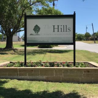 A welcoming sign for a location named 'Hills' surrounded by green grass and trees.