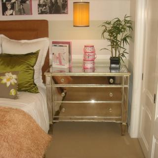 A cozy bedroom corner featuring a bed with pillows, a glass nightstand with decorative items, and a wall-mounted lamp.
