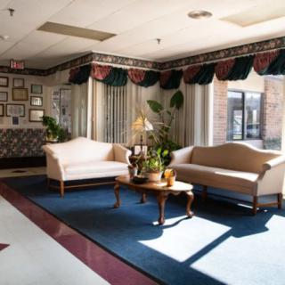 A well-lit lobby area with sofas and a table, decorated with plants and featuring large windows.