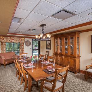 A well-furnished dining room with a long wooden table, chairs, and a china cabinet, adorned with a chandelier and large windows.