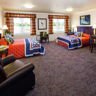 A cozy hotel room featuring two twin beds, colorful quilts, and a carpeted floor, illuminated by natural light from large windows.