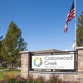 Entrance sign for Cottonwood Creek senior living community, featuring an American flag and surrounded by trees.