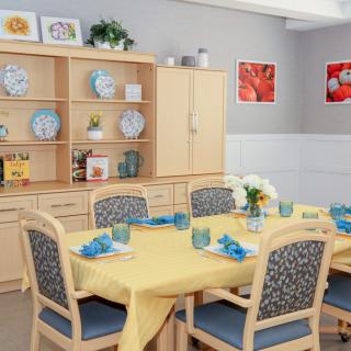 A neatly arranged dining room with a table set for four, featuring a yellow tablecloth and blue napkins, surrounded by wooden chairs and shelves displaying decorative plates.