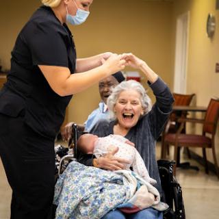 A heartwarming moment as a caregiver celebrates with an elderly woman holding a baby, showcasing the joy of connection and care.