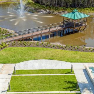 A scenic view of a park with a lake, fountain, and a gazebo connected by a bridge.