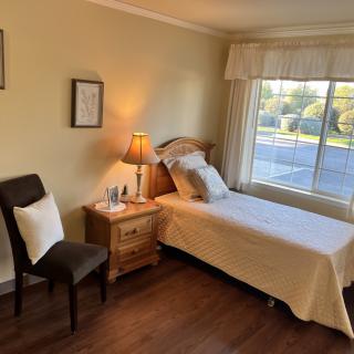 A cozy bedroom with a single bed, a nightstand with a lamp, a chair, and a large window letting in natural light.