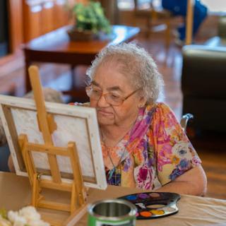 An elderly woman enjoying her time painting at an easel in a cozy indoor setting.